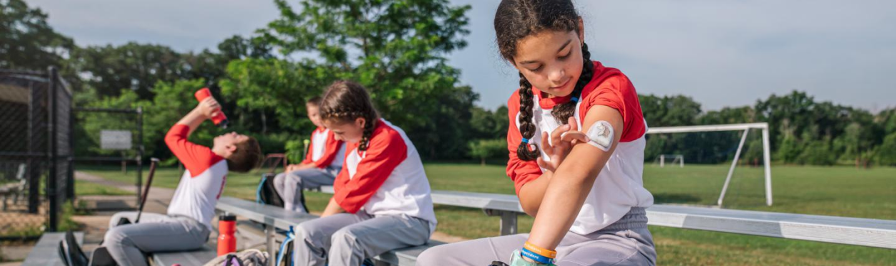 Youth baseball players on benches, one looking at the insulin pump on her arm